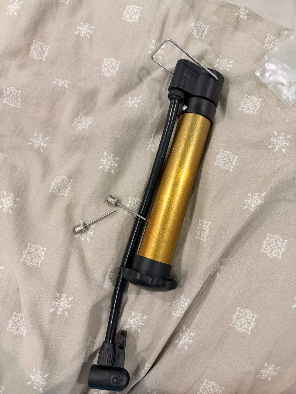 Black and gold-colored hand pump lying on a beige fabric with a floral pattern. The pump has a black handle and a