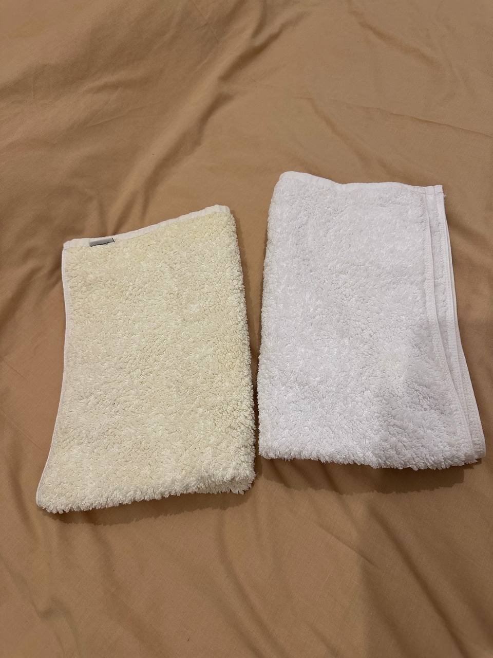 Two folded white towels on a beige-colored bedsheet. The towels appear to be made of a soft, fluffy material and have a