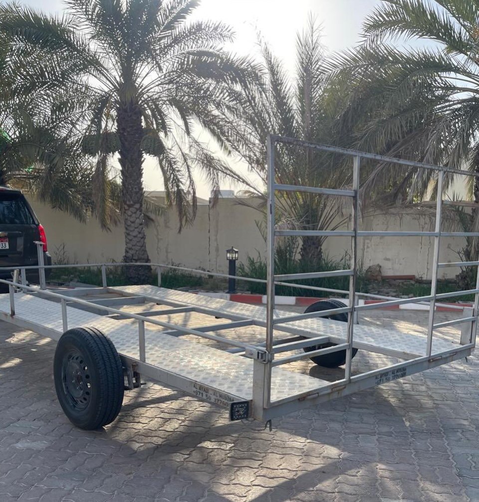 Metal trailer with a ramp attached to it. The trailer is parked on a cobblestone pavement with palm trees in the background.