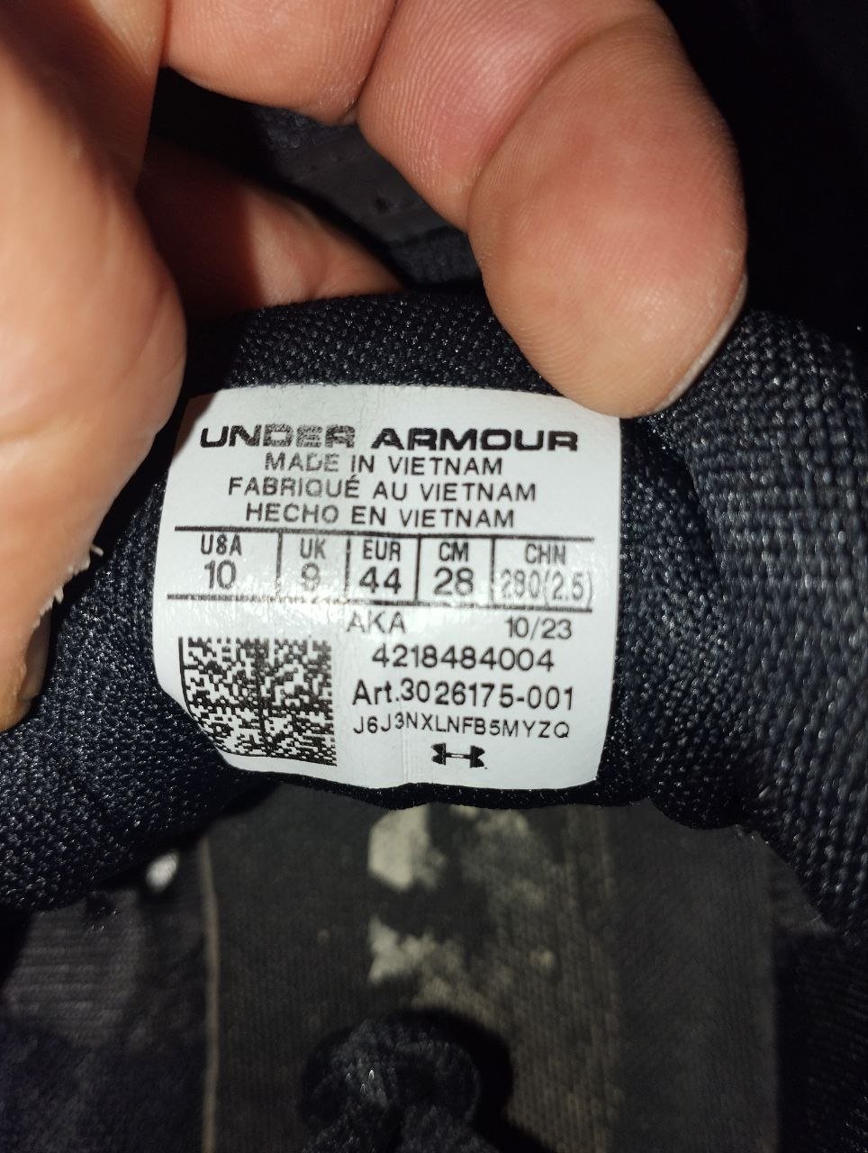 Close-up of a person's hand holding a black Under Armour label. The label is rectangular in shape and has white text and