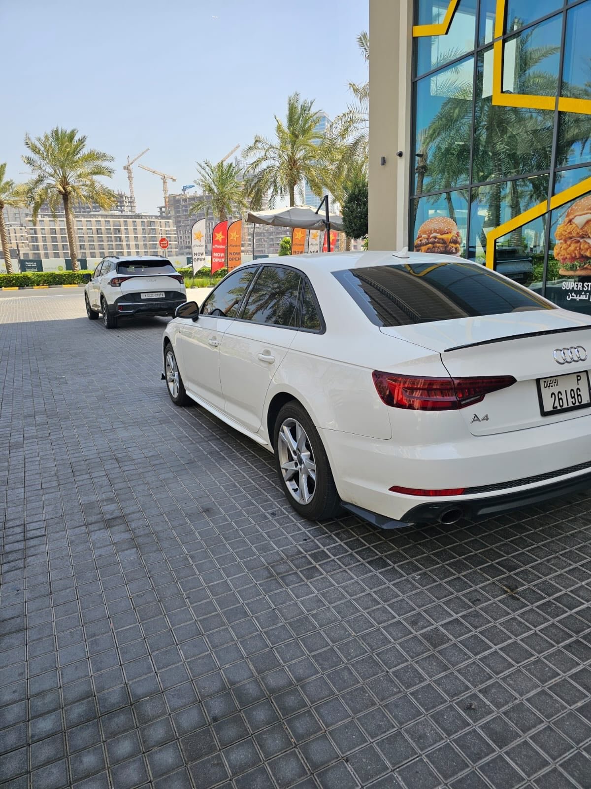 White Audi car parked in front of a McDonald's restaurant. The car is parked on a tiled pavement with palm trees in the