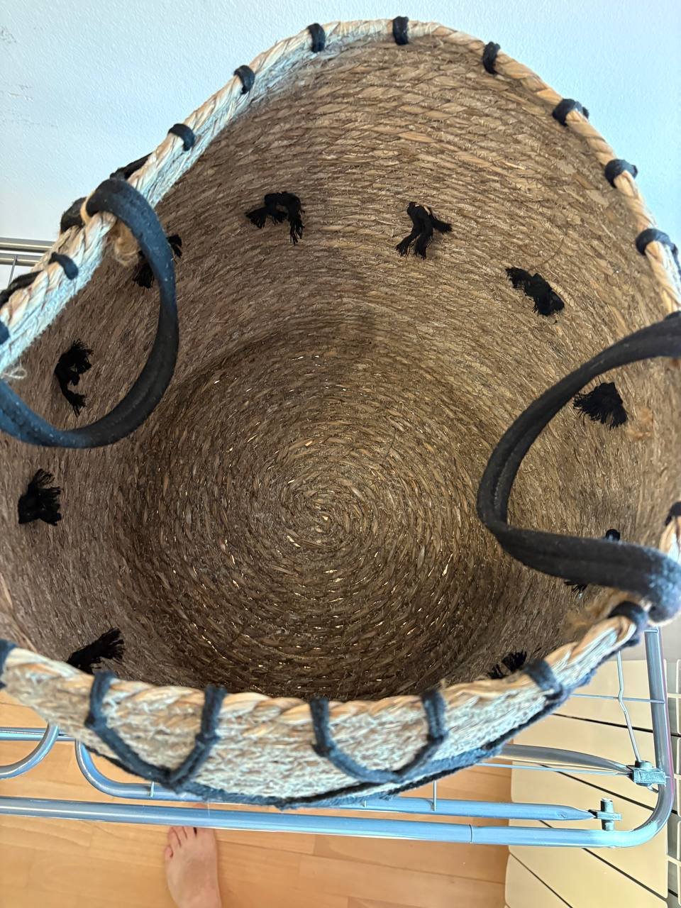 Round woven basket hanging on a metal rack. The basket is made of straw and has two black handles on either side. The