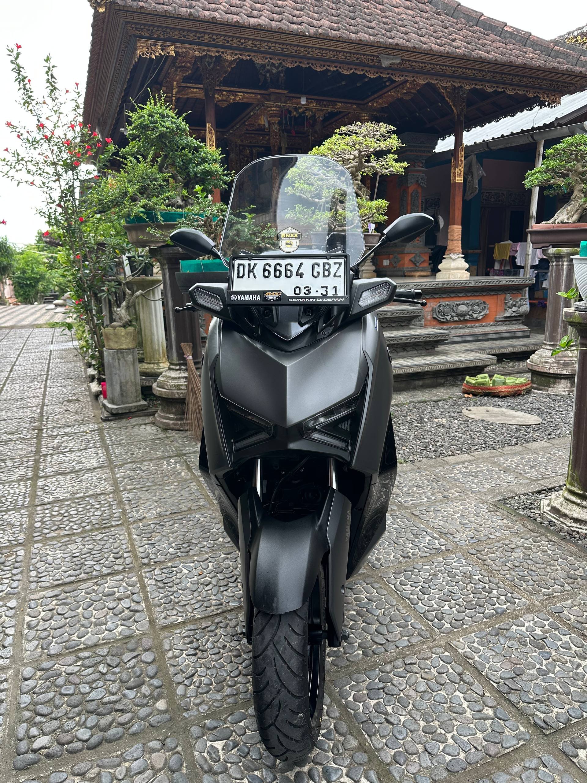 Black motorcycle parked on a cobblestone pavement in front of a traditional Thai temple. The temple has a thatched roof and
