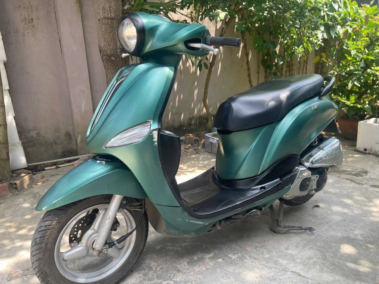 Green scooter parked on a concrete pavement. The scooter has a black seat and a round headlight on the front. The handlebars