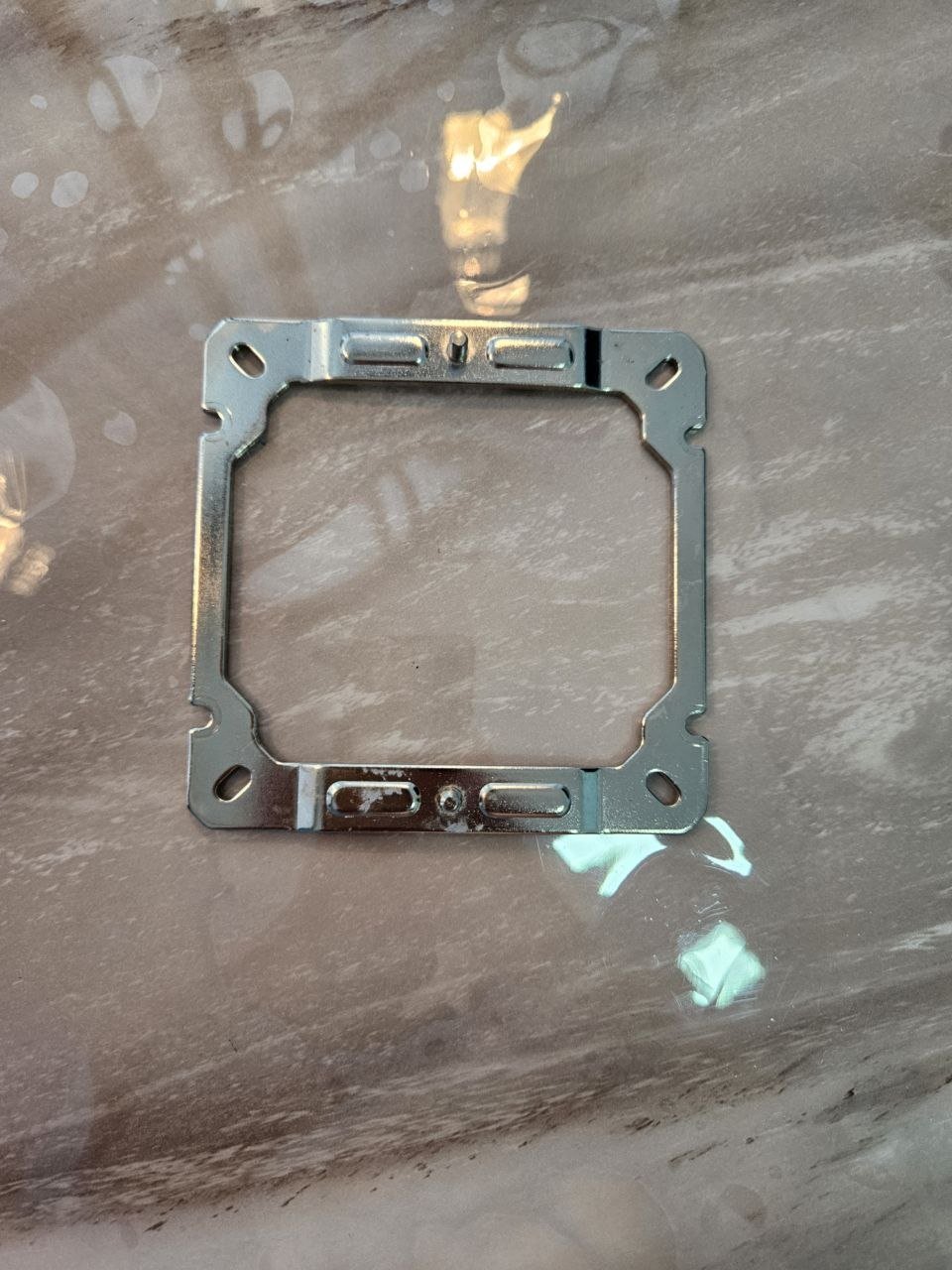Metal gasket or frame lying on a wooden surface. The gasket is rectangular in shape and appears to be made of metal with a